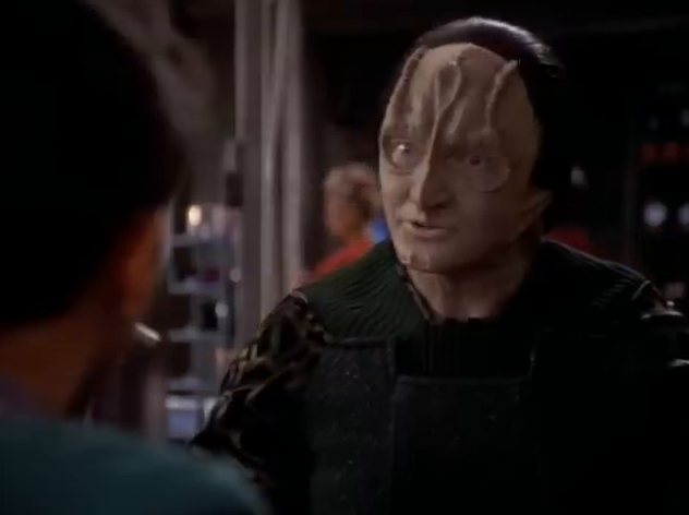 Garak, a Cardassian in a dark tunic, leaning across a table in a busy room.
