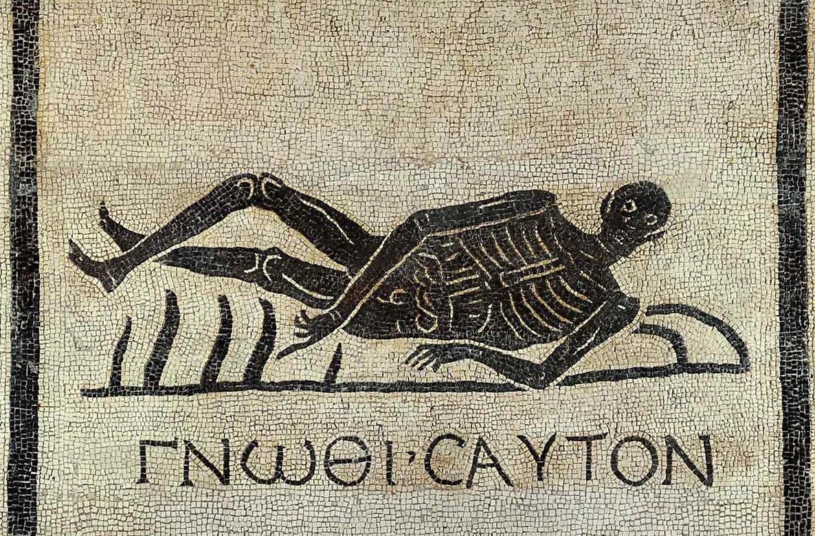 A black-and-white Roman floor mosaic of a skeleton lying on its side, with the Greek words for “know thyself” underneath.