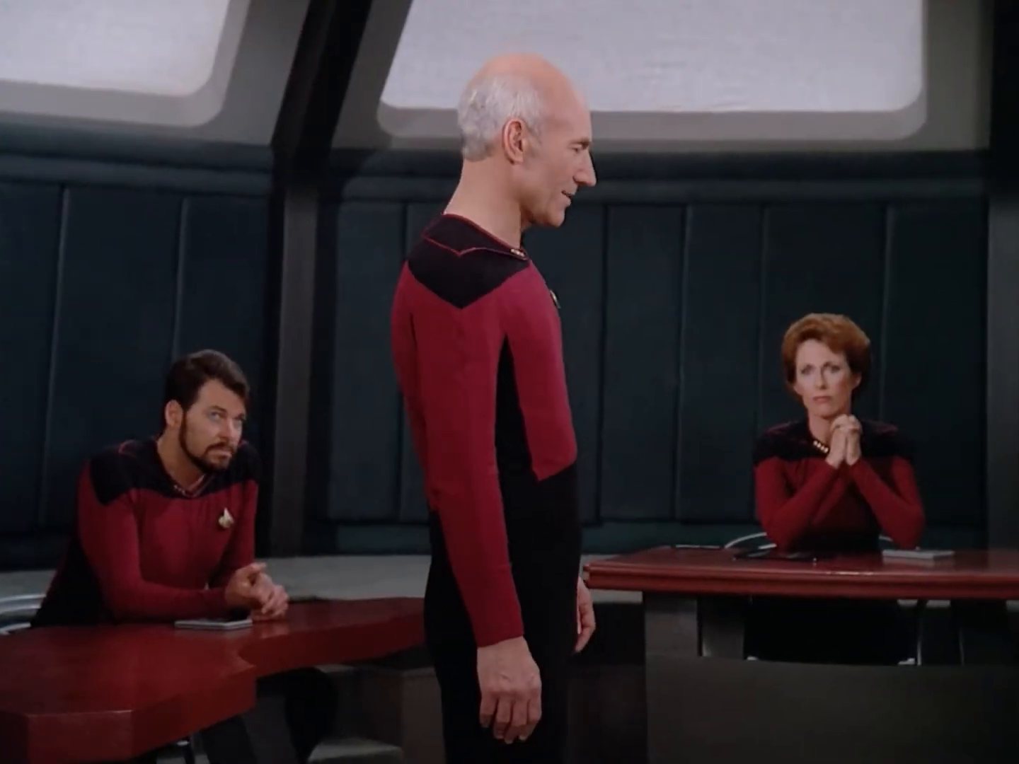 A small courtroom: Picard standing, Riker seated at left, the judge at right.