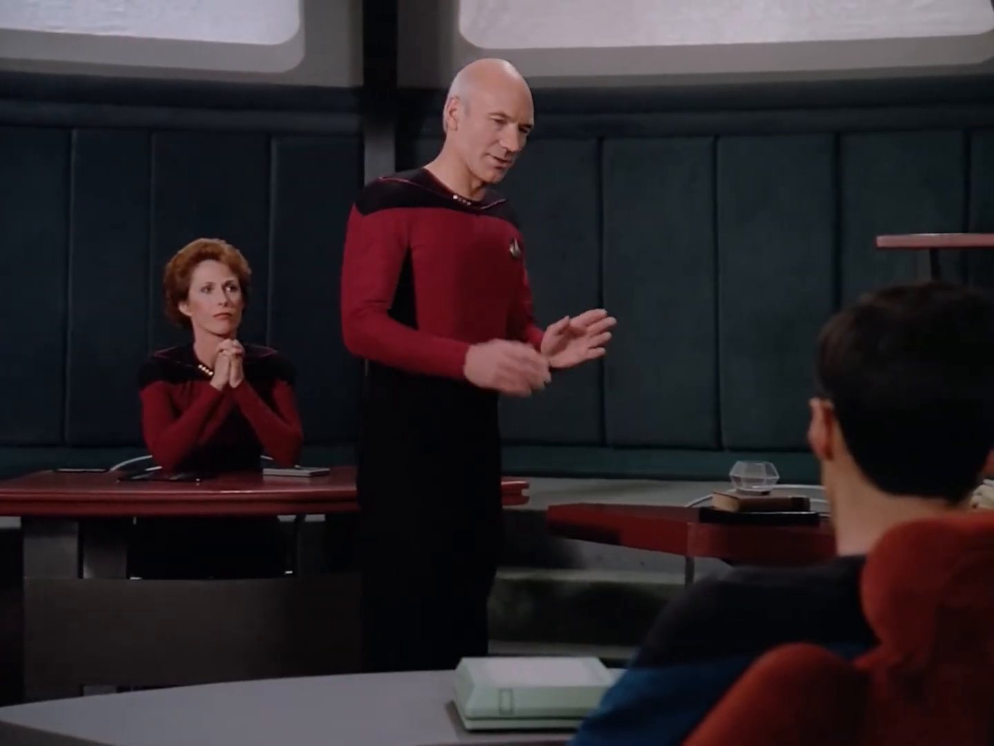 Data, an android in a gold uniform, seated at a table with his hands folded.