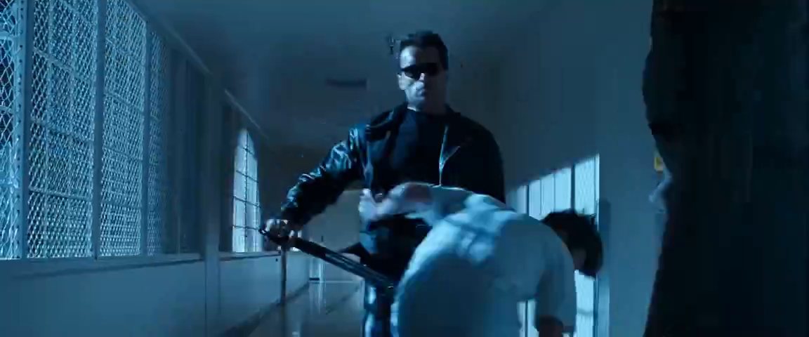 A man in dark glasses and a leather jacket, seen from below in a blue-lit corridor.