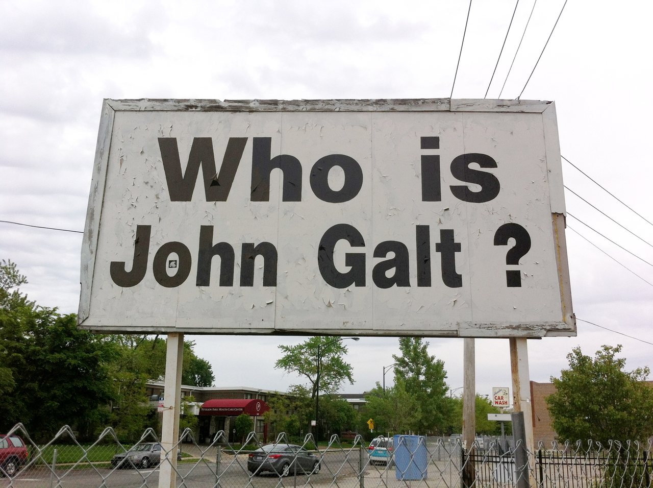 A weathered white billboard on posts, lettered in black: Who is John Galt? Trees, a fence and parked cars behind it.