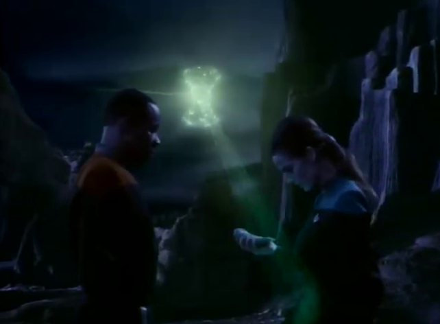 Two Starfleet officers on a dark, rocky plain, a glowing green shape hanging in the air behind them.