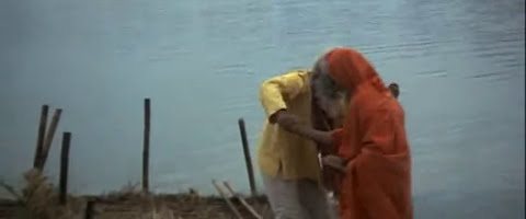 Two men on a riverbank: one in an orange robe leans in to kiss the forehead of the other, who wears yellow. Wide grey-blue water behind them.