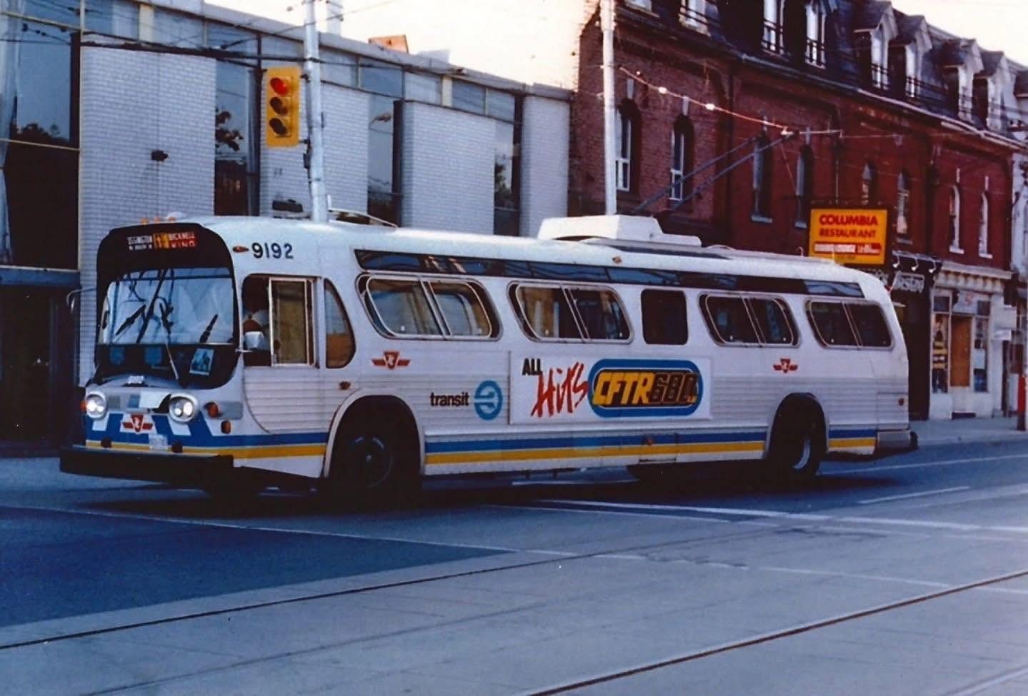 A white trolley bus with blue and yellow stripes, number 9192, crossing an intersection under overhead wires at dusk. An ad for CFTR 680 on its side; a brick row with a Columbia Restaurant sign behind it.