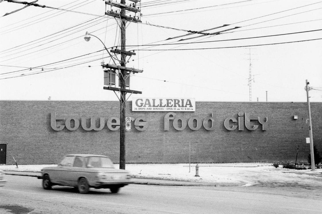 Black-and-white photo of a long brick wall with large letters reading towers and food city, a Galleria sign above them, and a car passing on the street.