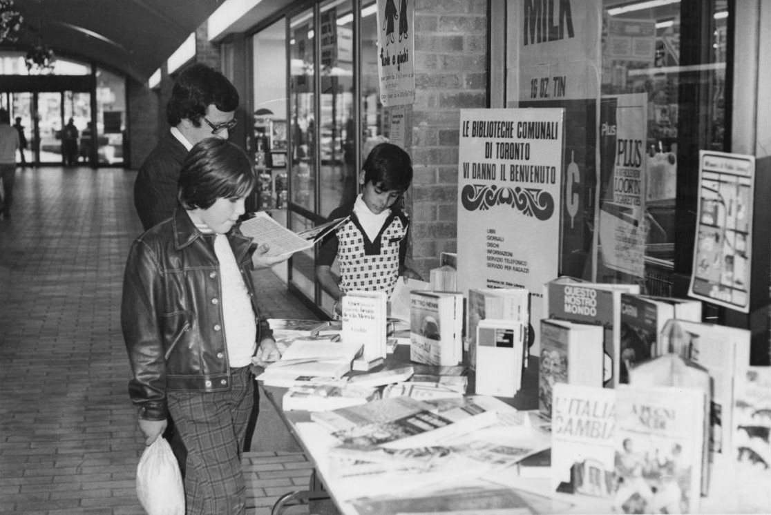 Black-and-white photo of two boys and a man looking over a table of Italian books in a mall corridor.